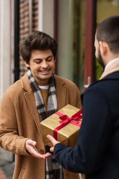 Pleased gay man in trendy clothes smiling near blurred boyfriend with Christmas present on city street — Stock Photo