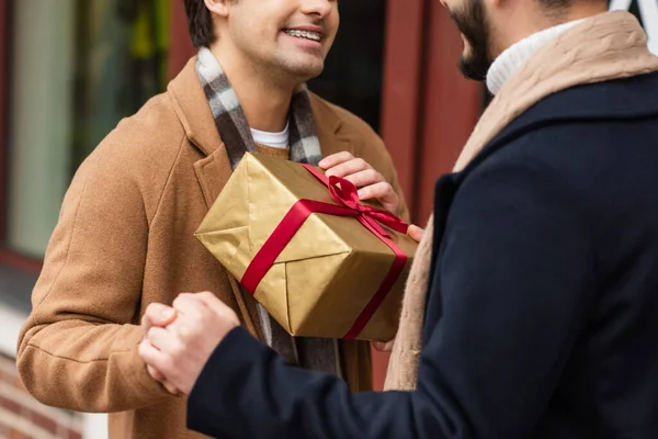 Visão parcial de sorrindo gay casal segurando as mãos perto de caixa de presente na rua turva — Fotografia de Stock