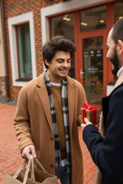Стильный гей с пакетами для покупок, принимающий рождественский подарок от парня на улице — стоковое фото