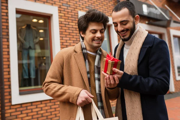 Young gay man with shopping bag holding gift box near happy bearded boyfriend and shop on blurred background — Stock Photo