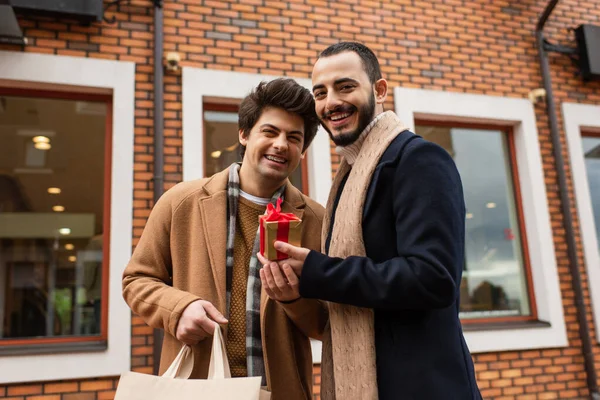 Alegre y elegante pareja gay sosteniendo bolsa de compras y regalo de Navidad cerca de tienda borrosa en la calle urbana - foto de stock