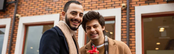 Feliz gay los hombres celebración pequeño regalo caja cerca tienda en borrosa fondo, bandera - foto de stock