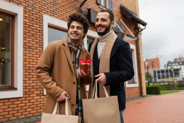 Cheerful and trendy gay men with Christmas present and shopping bags looking away near shop on urban street — Stock Photo