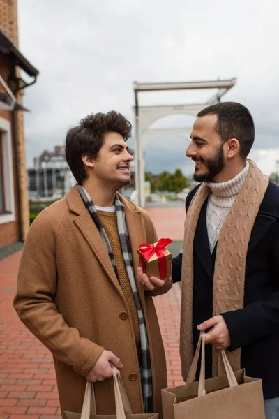 Young gay man with shopping bags and Christmas present looking at happy bearded boyfriend — Stock Photo