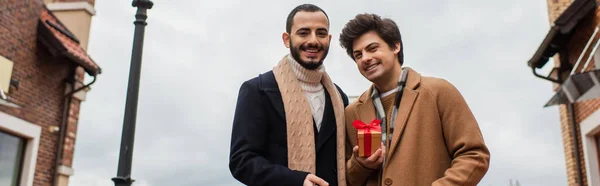 Молодые и счастливые геи смотрят в камеру рядом с маленькой подарочной коробкой на городской улице, баннер — стоковое фото