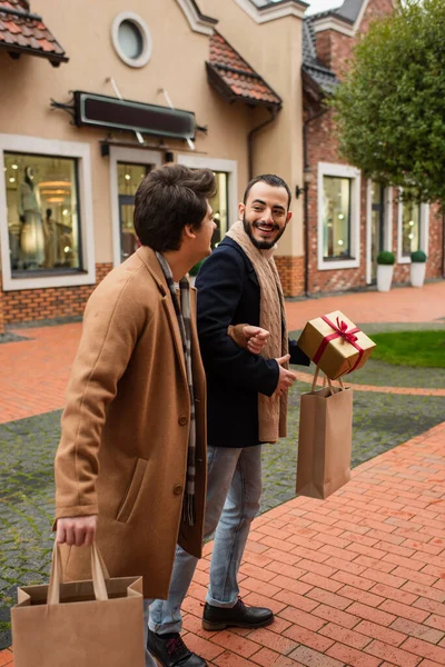 Щасливі геї з сумками для покупок та подарунковою коробкою, що йдуть по вулиці з розмитими магазинами — стокове фото