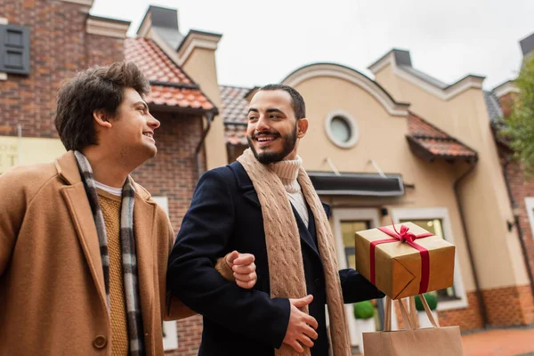 Bearded gay man with shopping bag and Christmas present looking at young boyfriend on urban street — Stock Photo