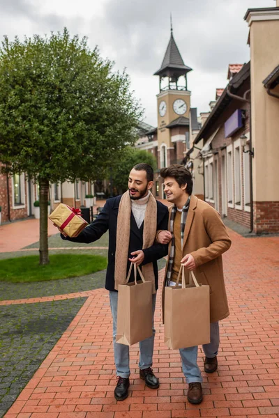 Comprimento total de parceiros gays na moda com sacos de compras olhando para caixa de presente na rua urbana — Fotografia de Stock