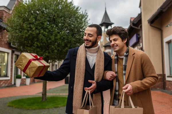 Excité et à la mode gay couple tenant des sacs à provisions et regardant boîte cadeau sur la rue — Photo de stock