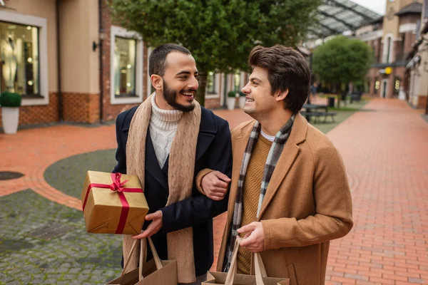 Happy gay couple in stylish clothes holding gift box and shopping bags while smiling at each other on street — Stock Photo
