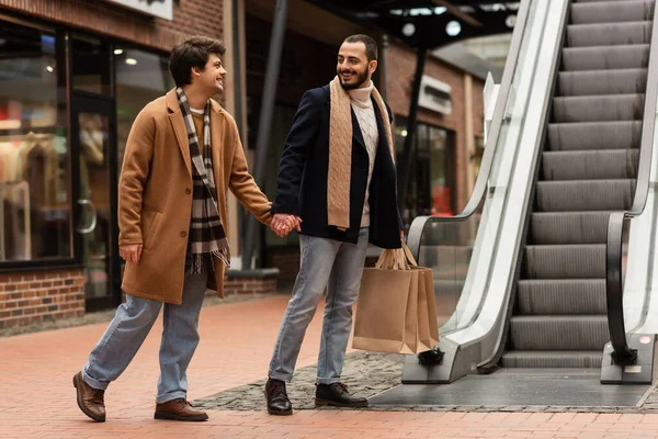 Full length of bearded gay man with shopping bags holding hands with happy boyfriend near escalator — Stock Photo