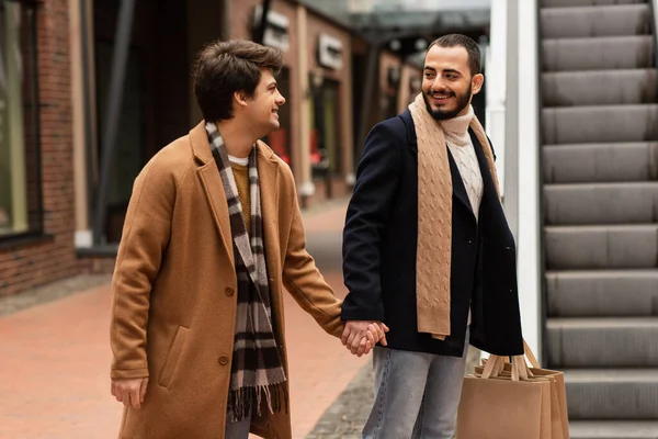 Happy gay man with shopping bags holding hands with stylish boyfriend near blurred escalator — Stock Photo