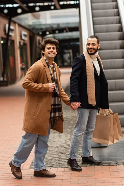 Gay hombre en beige abrigo y a cuadros bufanda cogido de la mano con sonriente barbudo novio con compras bolsas cerca de escaleras mecánicas - foto de stock