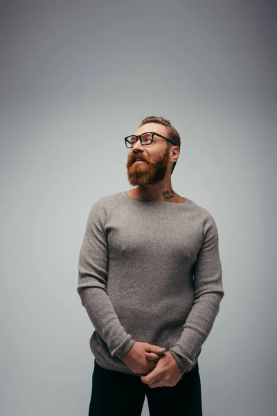 Sorridente uomo tatuato in occhiali e maglione guardando lontano isolato sul grigio — Foto stock