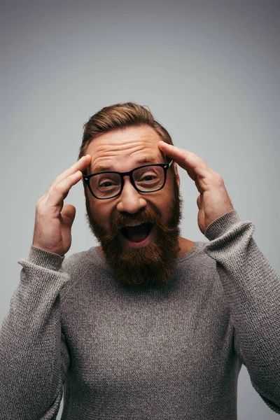 Волнующий и бородатый мужчина в очках смотрит на камеру, изолированную на сером — стоковое фото