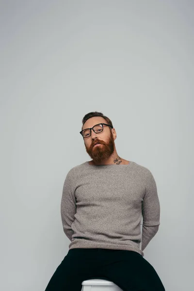 Модний бородатий чоловік в окулярах позує на стільці ізольовано на сірому — стокове фото
