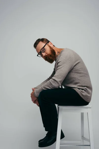Стильный мужчина в джинсах и очках, сидящий на стуле, изолированный на сером — стоковое фото
