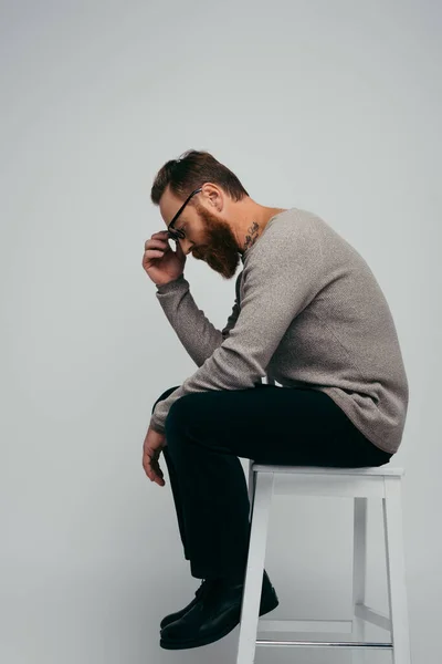 Seitenansicht eines bärtigen Mannes, der eine Brille berührt, während er auf einem Stuhl sitzt, isoliert auf grau — Stockfoto