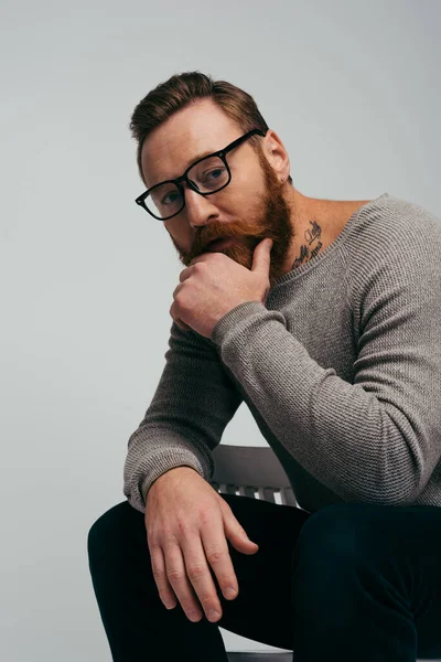 Porträt eines bärtigen Mannes mit Brille, der sein Kinn berührt, während er isoliert auf einem Stuhl in grau sitzt — Stockfoto