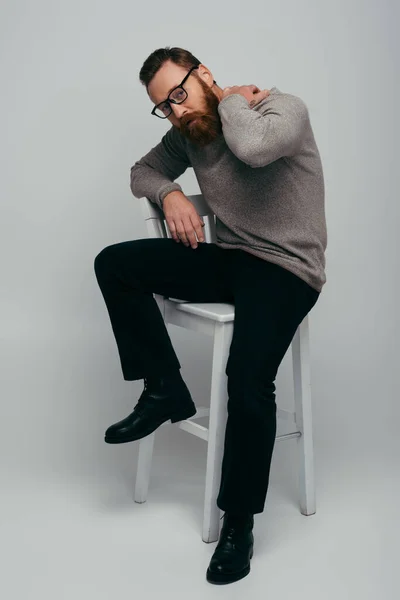 Longitud completa del hombre barbudo de moda tocando el cuello mientras está sentado en la silla sobre fondo gris - foto de stock