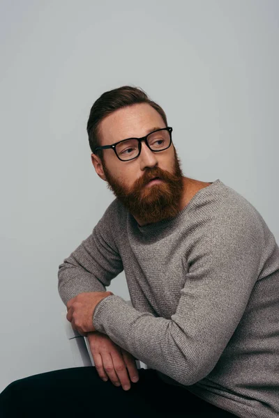 Modelo de moda en gafas y jersey sentado en silla aislada en gris - foto de stock