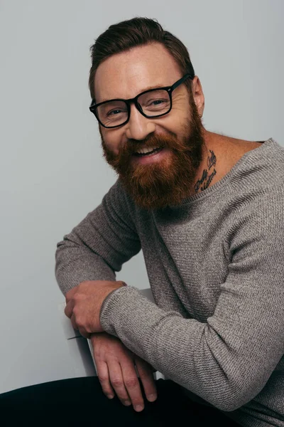 Портрет позитивного человека в очках и свитере, сидящего на стуле изолированно на сером — стоковое фото