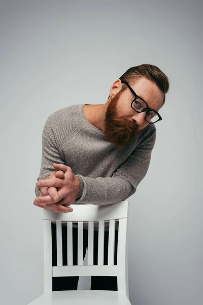 El hombre a la moda en las gafas que miran la cámara cerca de la silla aislada en gris - foto de stock
