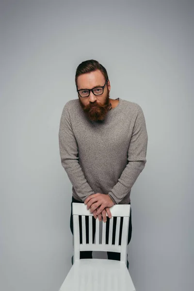 Бородатый мужчина в свитере смотрит в камеру рядом с белым стулом, изолированным на сером — стоковое фото