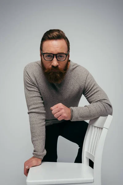 Bärtiger Mann mit Brille blickt in die Kamera, während er in der Nähe von Stuhl isoliert auf grau posiert — Stockfoto