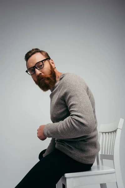 Стильный бородатый мужчина в очках, сидящий на белом стуле, изолированном на сером — стоковое фото