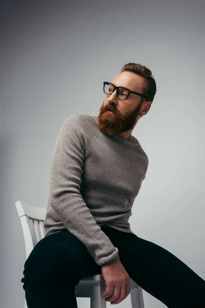 Модний бородатий чоловік у джемпер позує на стільці на сірому фоні — стокове фото