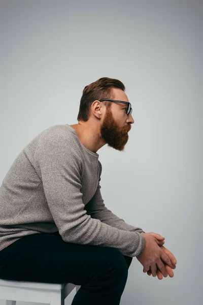 Seitenansicht eines bärtigen Mannes mit Brille, der wegschaut, während er isoliert auf einem Stuhl in grau sitzt — Stockfoto