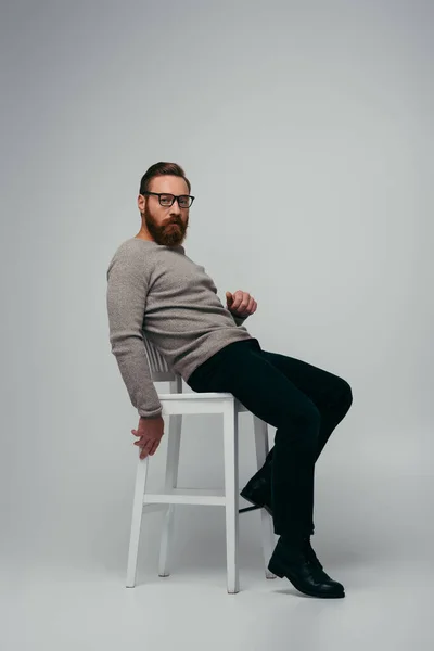 Full length of trendy bearded man in eyeglasses looking at camera while sitting on chair on grey background — Stock Photo