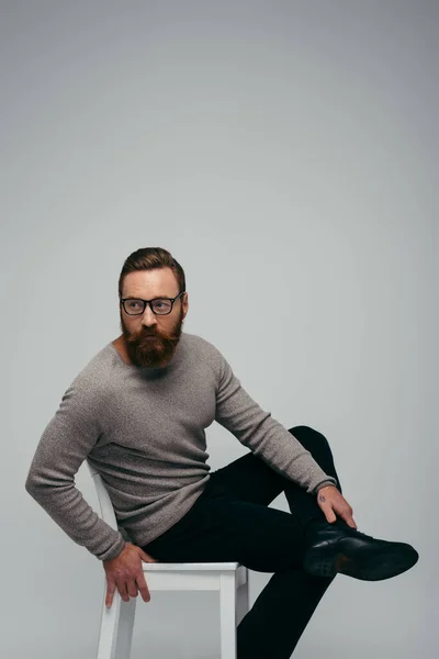 Fashionable model in eyeglasses looking away while sitting on chair isolated on grey — Stock Photo
