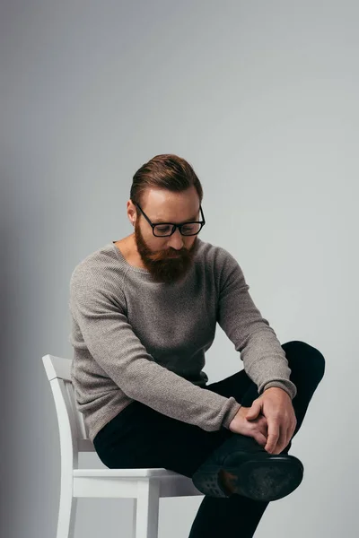 Homme barbu à la mode dans des lunettes de vue toucher la jambe tout en étant assis sur une chaise sur fond gris — Photo de stock