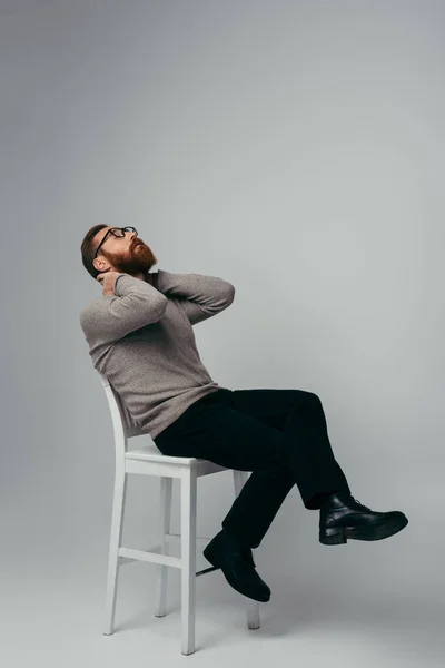 Lunghezza intera del modello in maglione e occhiali toccando il collo mentre seduto su una sedia su sfondo grigio — Foto stock