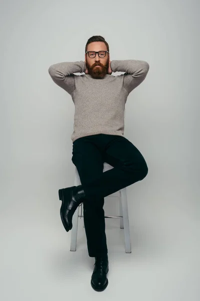 Comprimento total de homem elegante em óculos tocando no pescoço e olhando para a câmera enquanto sentado na cadeira em fundo cinza — Fotografia de Stock