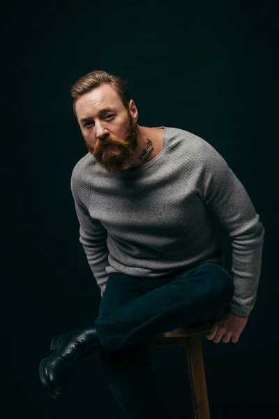 Тенді-бородатий чоловік у сірому светрі сидить на стільці і дивиться на камеру, ізольовану на чорному — стокове фото