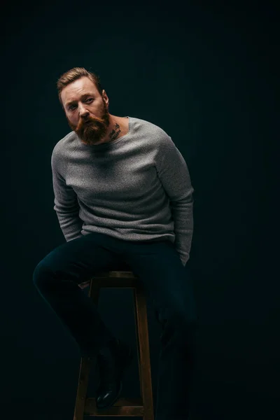 Bearded man in stylish grey jumper sitting on chair isolated on black — Stock Photo