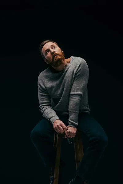 Bearded man in jumper holding eyeglasses and looking up while sitting on chair isolated on black — Stock Photo