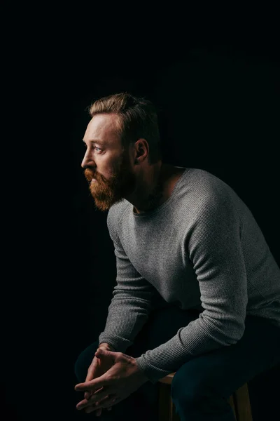 Бородатый мужчина в сером свитере смотрит в сторону, изолированный от тьмы — стоковое фото