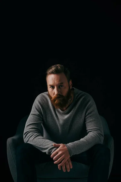 Modischer Mann im Pullover blickt in die Kamera in der Nähe von Sessel isoliert auf schwarz — Stockfoto