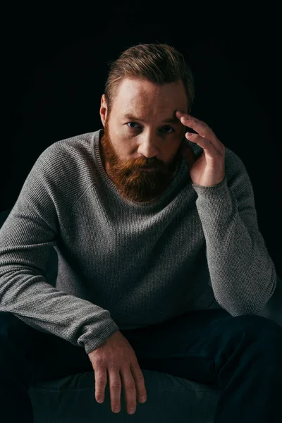 Портрет бородатого мужчины, смотрящего в камеру возле кресла, изолированного на черном — стоковое фото