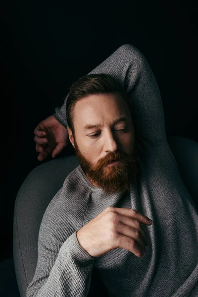 Stylish bearded man in jumper sitting on armchair isolated on black with shadow — Stock Photo