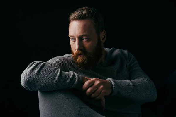 Bearded model in grey jumper looking away while sitting on armchair isolated on black — Stock Photo