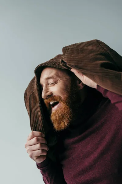 Hombre barbudo con chaqueta marrón en la cabeza sonriendo aislado en gris - foto de stock