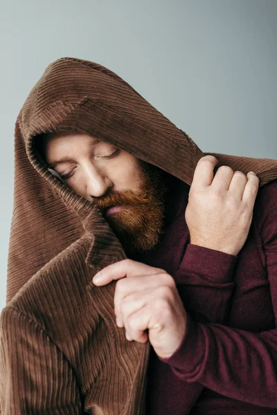 Бородатый мужчина с коричневым блейзером на голове и закрытыми глазами позирует изолированно на сером — стоковое фото