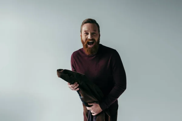 Excited and bearded man in turtleneck holding blazer while looking at camera isolated on grey — Stock Photo