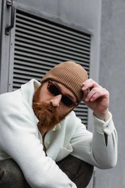 Бородатый мужчина в стильных солнцезащитных очках и модной шляпе-шапочке, смотрящий на улицу — стоковое фото