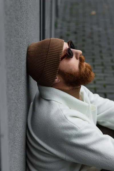 Side view of bearded man in stylish sunglasses and trendy beanie hat leaning on grey wall — Stock Photo